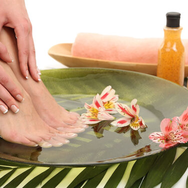 Feet soaking in a bowl with pink flowers, surrounded by wellness items like a towel, a bottle, and leaves, symbolizing the benefits of an Aqua-Ionic Detox Foot Spa.