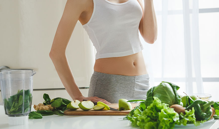 Person in white tank top preparing a healthy green smoothie with fresh vegetables and fruits on a kitchen counter.