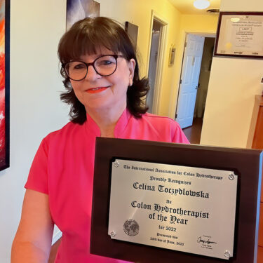 Celina Toczydlowska in a pink top holds a plaque recognizing her as Colon Hydrotherapist of the Year 2022 in an office setting.