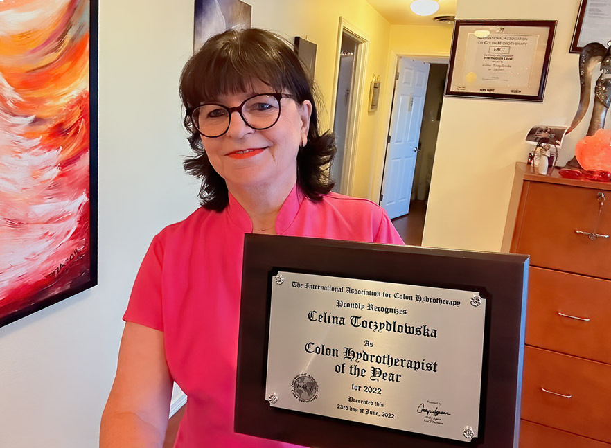 Celina Toczydlowska in a pink top holds a plaque recognizing her as Colon Hydrotherapist of the Year 2022 in an office setting.