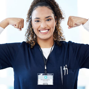 Young healthcare professional in a blue uniform, smiling and flexing arms, wearing a medical ID badge.