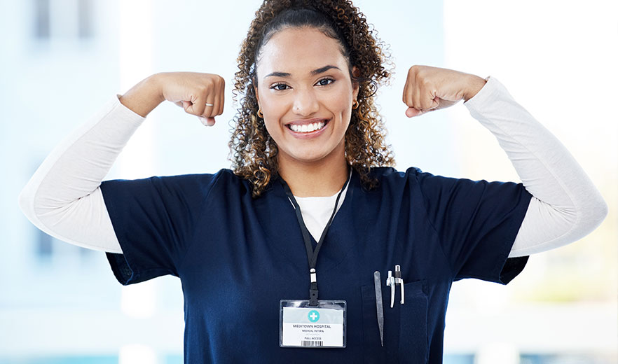 Young healthcare professional in a blue uniform, smiling and flexing arms, wearing a medical ID badge.