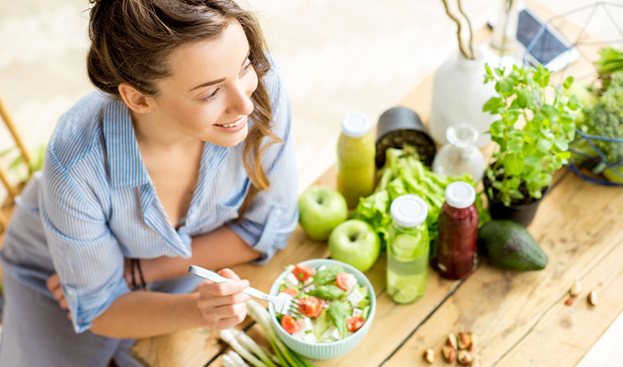 Woman enjoying a fresh salad surrounded by fruits, vegetables, and juices on a wooden table, promoting healthy living and wellness.