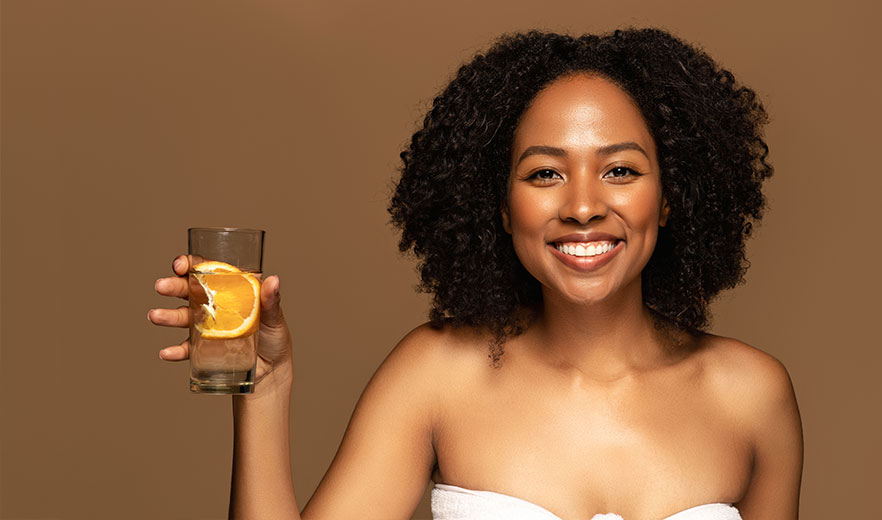 Smiling woman holding a glass of water with orange slices, wearing a towel, symbolizing body cleansing and colon hydrotherapy.