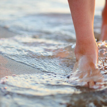 Feet walking on wet sand at the beach, with gentle waves washing over them.