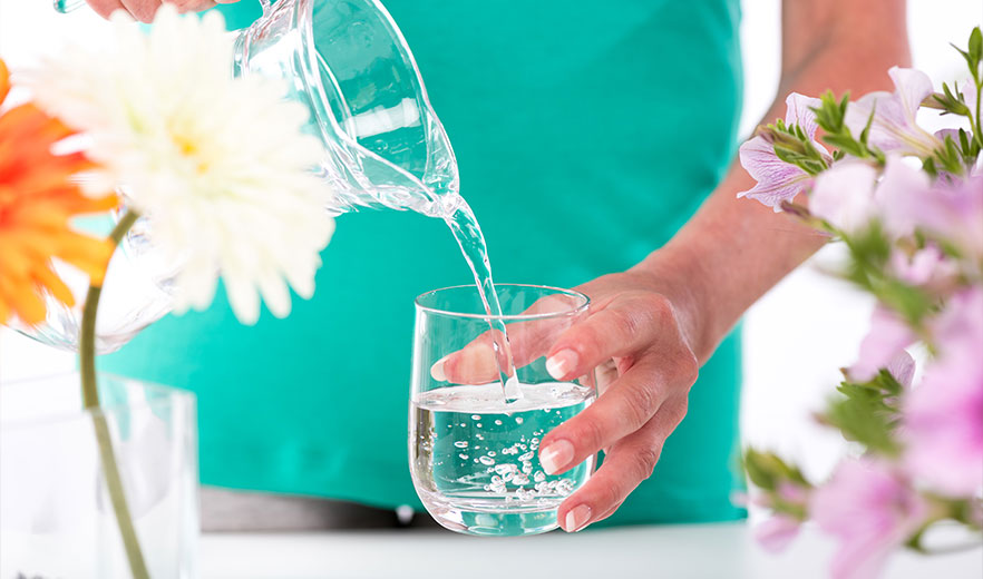 Person in turquoise shirt pours water into a glass, surrounded by colorful flowers, promoting hydration and wellness.