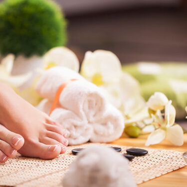 Feet resting on a spa mat, surrounded by orchids, rolled towels, and stones, conveying a tranquil spa atmosphere for relaxation.