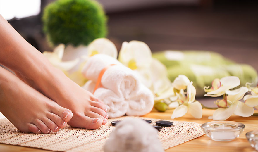 Feet resting on a spa mat, surrounded by orchids, rolled towels, and stones, conveying a tranquil spa atmosphere for relaxation.