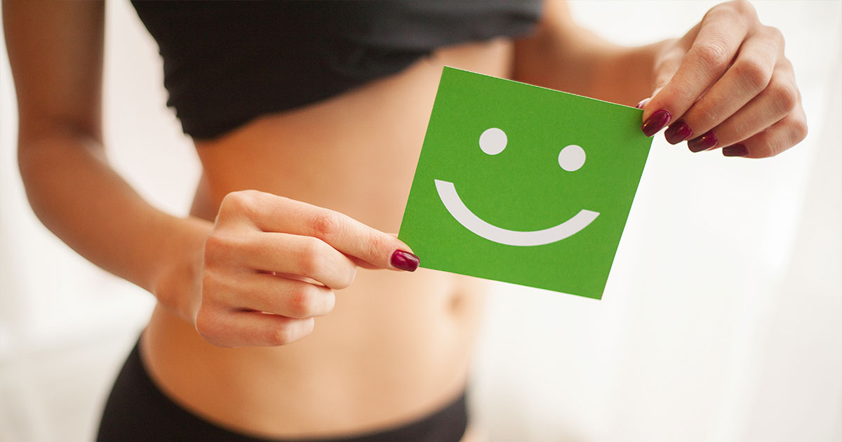 Person holding a green smiley face card in front of stomach, promoting digestive wellness and colon hydrotherapy benefits.