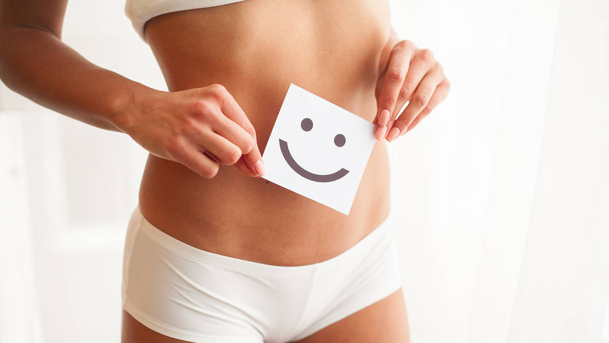 Smiling woman holding a happy face card over her abdomen, symbolizing digestive wellness after colon hydrotherapy at a wellness center in Bellevue.