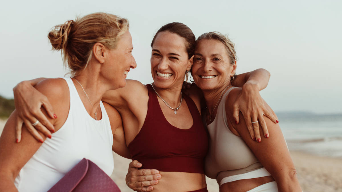 Three smiling women at the beach embracing after a wellness session, promoting healthy living and community at a wellness center in Bellevue.