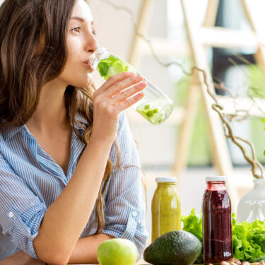 Woman drinking infused water beside fresh greens and juices, illustrating hydration tips for constipation and colon hydrotherapy support at ERC Bellevue.