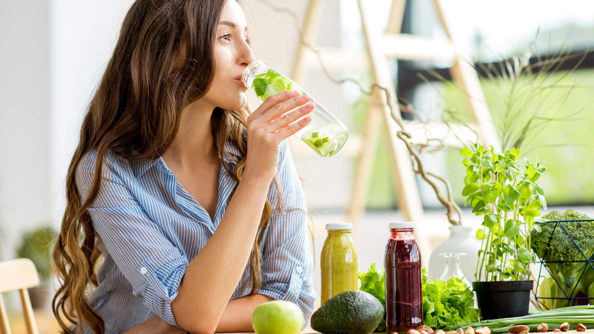 Woman drinking infused water beside fresh greens and juices, illustrating hydration tips for constipation and colon hydrotherapy support at ERC Bellevue.