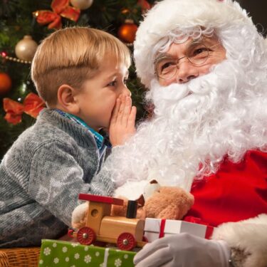 A child whispers to Santa Claus beside a decorated Christmas tree, holding a toy train and gifts.