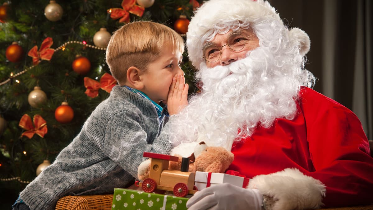 A child whispers to Santa Claus beside a decorated Christmas tree, holding a toy train and gifts.