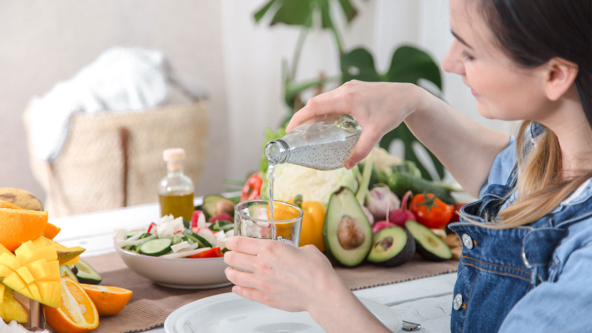 Woman pouring water into a glass beside fresh fruit and vegetables, supporting hydration habits for bloating and water retention.